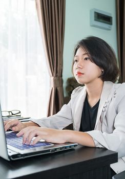 Young asian woman short hair in smart casual wear working on laptop while sitting near window in home office