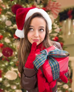 Thinking Girl Wearing A Christmas Santa Hat with Bow Wrapped Gift In Front of Decorated Tree.