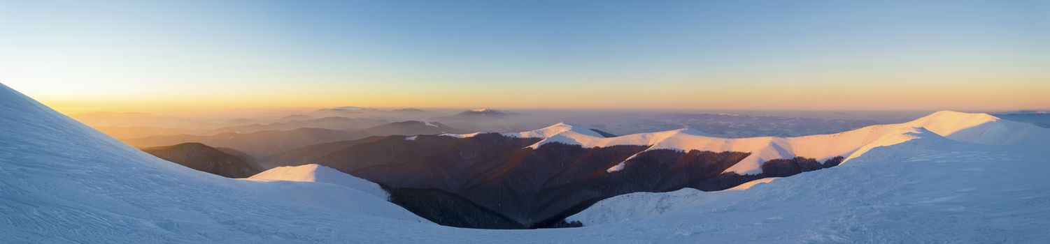 Wide panorama of winter sunset in the mountains. Gentle sun rays enlighten snowy mountain ridge. Traces of wind and frost in the snow. Ukrainian Carpathians