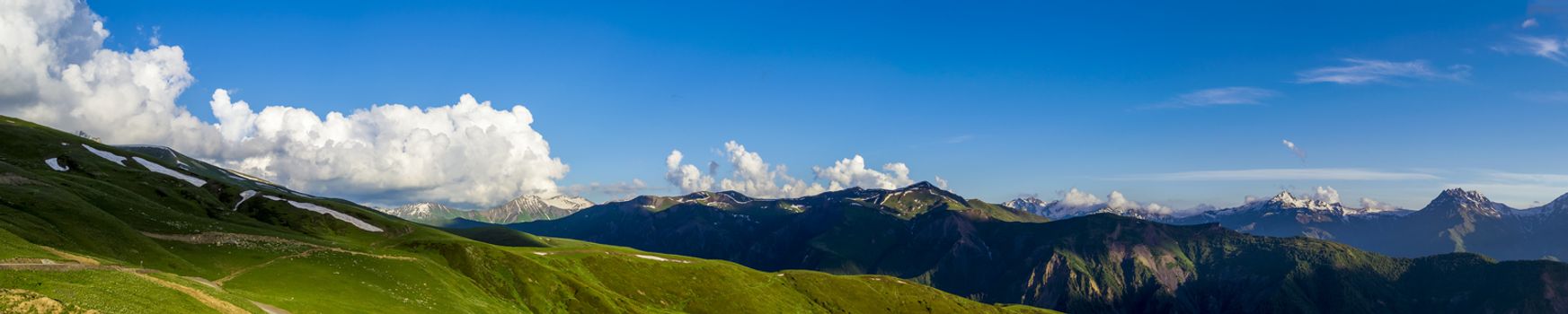 Wide mountain panorama.Green hills with snow on peaks in evening sunlight. Dark shadows. Blue sky with some clouds. Summer in Georgia.