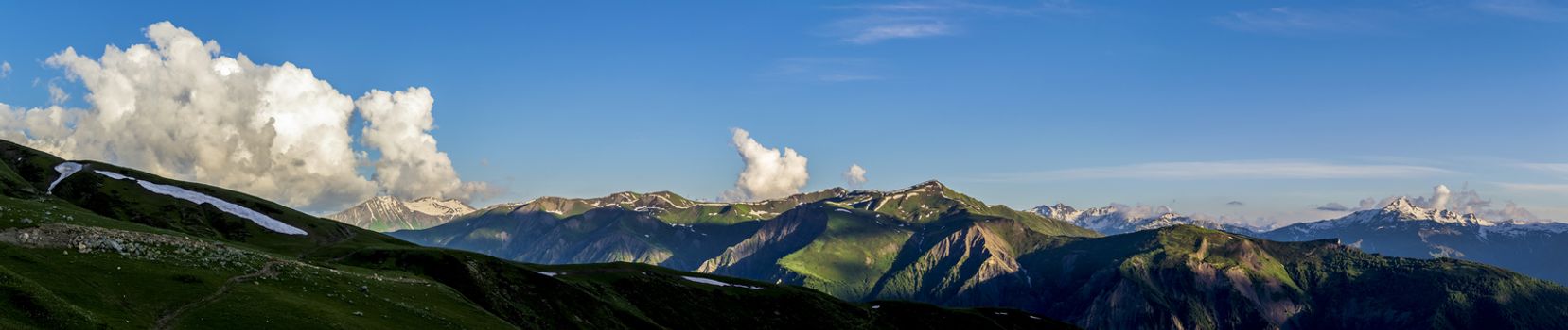 Wide mountain panorama.Green hills with snow on peaks in evening sunlight. Dark shadows. Blue sky with some clouds. Summer in Georgia.