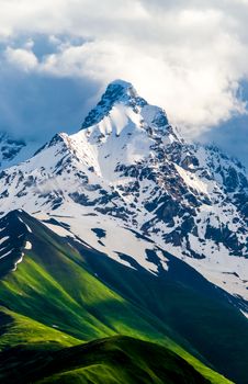 Images of high and snow-covered mountain peak. White clouds are visible behind the lighted mountain top. Summer in Caucasian Georgia. Vertical orientation photo.