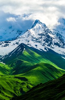 Beautiful view of the snowy mountain peak. Green fields and hills changes to snow. Storm white clouds are visible behind the lighted mountain top. Summer in Georgia. Caucasus. Svaneti, Ushguli. Vertical orientation photo.