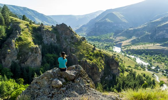 Girl sitting on a rock looking at the view. Against the background of sharp rocks, hills, valley. Clear sky, green hills, steep cliffs, wide river - the extraordinary nature of Georgia.