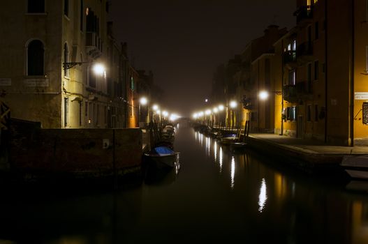 Quiet canal at night in Venice. Warm light of the street lights illuminates the street and reflected in waterways. Private boats are parked along the canal. The bridge silhouette is visible at the end of the waterway