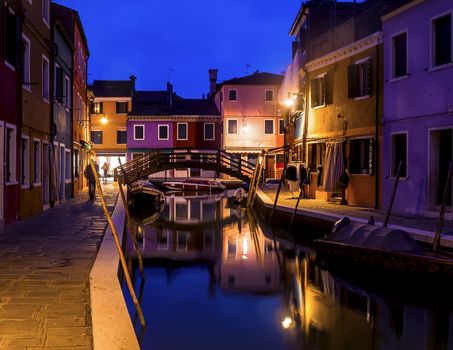 Evening cityscape of Burano island in Venice. Colored buildings and a small bridge illuminated by street lights, reflected in a water canal.