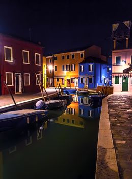 Beautiful night cityscape. Colored buildings are illuminated by street lights reflected in a water canal. Boats are parked there. Venice, Burano island.