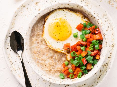 Savory oatmeal in craft trendy bowl, served with vegetables and fried egg on white concrete background. Red bell pepper and bunch onion on background. Top view or flat lay.