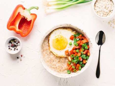 Savory oatmeal in craft trendy bowl, served with vegetables and fried egg on white concrete background. Red bell pepper and bunch onion on background. Top view or flat lay. Copy space.