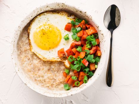 Savory oatmeal in craft trendy bowl, served with vegetables and fried egg on white concrete background. Red bell pepper and bunch onion on background. Top view or flat lay.