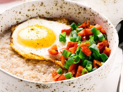 Savory oatmeal in craft trendy bowl, served with vegetables and fried egg on white concrete background. Red bell pepper and bunch onion on background.