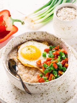 Savory oatmeal in craft trendy bowl, served with vegetables and fried egg on white concrete background. Red bell pepper and bunch onion on background. Vertical