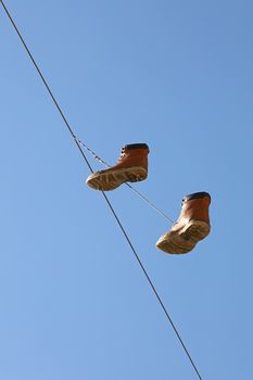 Pair of old worn brown boots hanging on a telephone line wire as traditional urban youth joke, over clear blue sky, low angle view