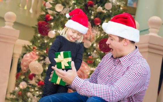 Happy Young Girl and Father Wearing Santa Hats Opening Gift Box In Front of Decorated Christmas Tree.