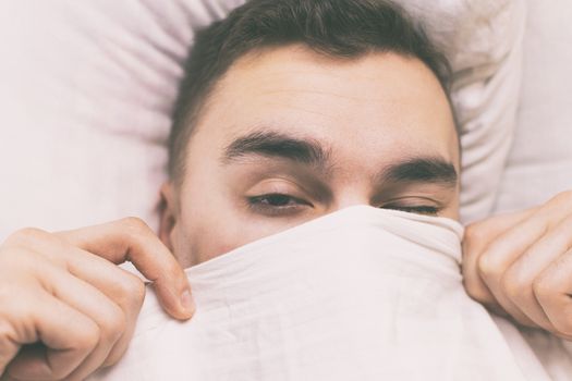 A young man wakes up in the morning under a blanket.