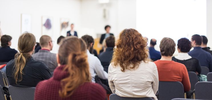 Business and entrepreneurship symposium. Female speaker giving a talk at business meeting. Audience in conference hall. Rear view of unrecognized participant in audience. Copy space on whitescreen.