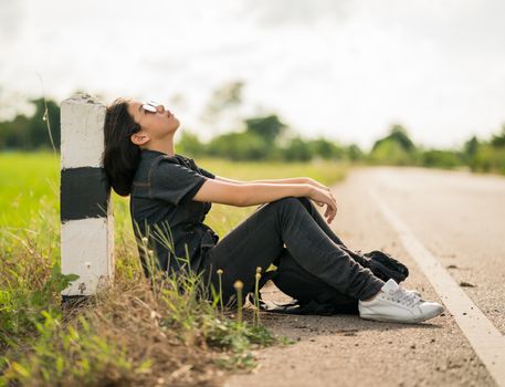 Young asian woman short hair and wearing sunglasses sit with backpack hitchhiking along a road wait for help in country road Thailand