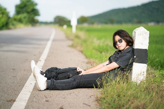 Young asian woman short hair and wearing sunglasses sit with backpack hitchhiking along a road wait for help in country road Thailand