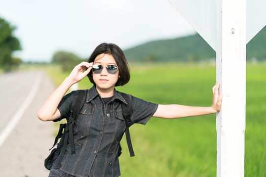 Young asian woman short hair and wearing sunglasses with backpack hitchhiking along a road in countryside Thailand