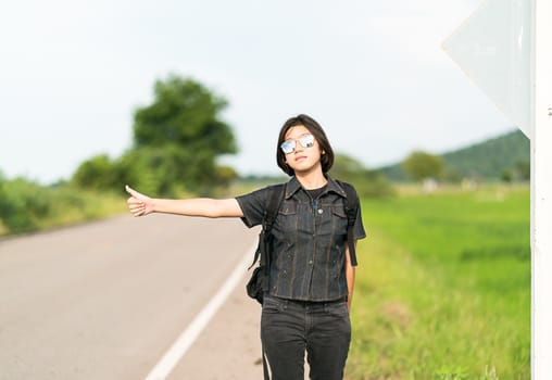 Young asian woman short hair and wearing sunglasses with backpack hitchhiking along a road in countryside Thailand