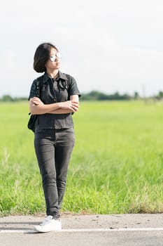 Young asian woman short hair and wearing sunglasses with backpack hitchhiking along a road in countryside Thailand