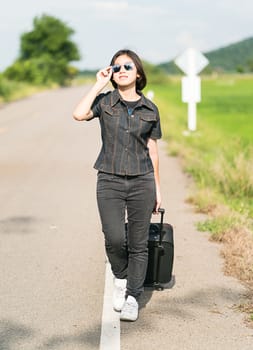 Young asian woman short hair and wearing sunglasses with luggage hitchhiking along a road in countryside Thailand