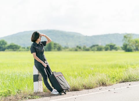 Young asian woman short hair and wearing sunglasses with luggage hitchhiking along a road in countryside Thailand