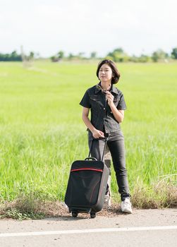 Young asian woman short hair and wearing sunglasses with luggage hitchhiking along a road in countryside Thailand