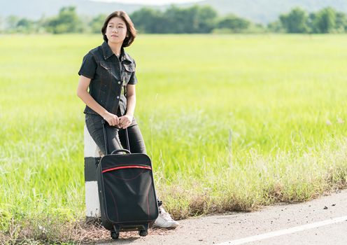 Young asian woman short hair and wearing sunglasses with luggage hitchhiking along a road in countryside Thailand