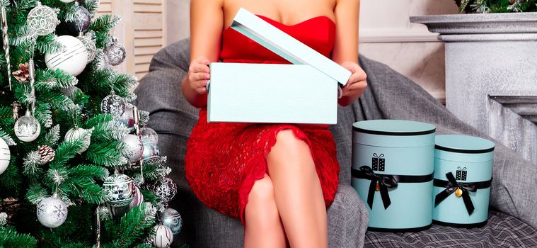 Woman in red dress opening her christmas gift boxes while sitting at home near by christmas tree. Holidays and celebration concept