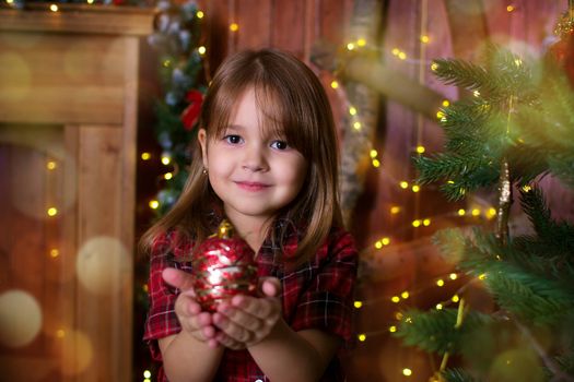 The girl holds in hand a Christmas ball near a Christmas tree