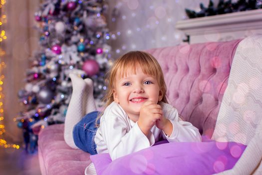 The little girl lies on a sofa against the background of a Christmas tree in a photographic studio