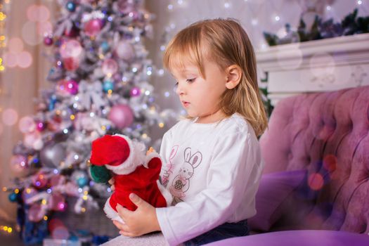 the little girl sits on a sofa near a Christmas tree, and holds toy Santa Claus in hand