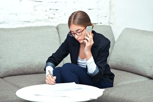 Beautiful young business woman sitting on sofa at office or at home, calling on phone and working with documents