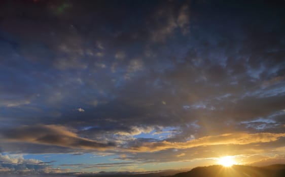 Sun setting sunburst behind mountain with blue sky and clouds tinged with colour.  The image shows primarily sky