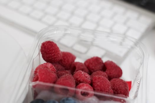 Fresh and tasty raspberries near white computer keyboard in office.