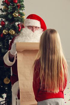 Santa Claus with with list or nice or naugthy list paper with small girl near christmas tree