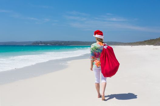 Christmas for a lot of Australians means hot summer sunshine and days spent at the beach.  A woman wearing a santa hat strolls along a white sandy beach with a red drawstring sack.   Space for copy 