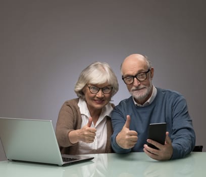 Senior couple with laptop and smartphone showing thumbs up