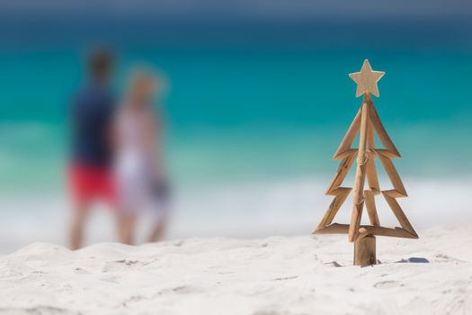 Rustic timber driftwood Christmas tree with star on a white sandy beach with out o f focus ocean snd indistinguishable people strolling by