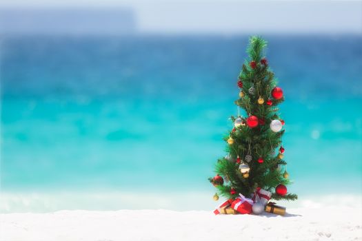 Christmas tree decorated with colourful baubles and presents underneath it, stands on a beautiful sandy beach with background blur of the ocean and sky. 
