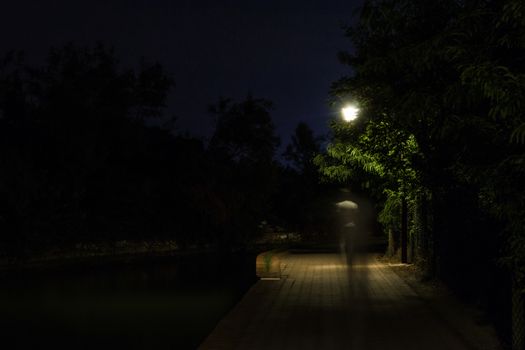 Double exposure night scene of person walking dark street illuminated with streetlights. The receding male silhouettes on the road in the park. Human figure in motion blur going along the city river. Long shutter.