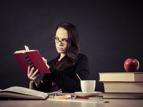 Photo of a teacher in her 30's sitting at a messy desk in front of a large blackboard reading a book.