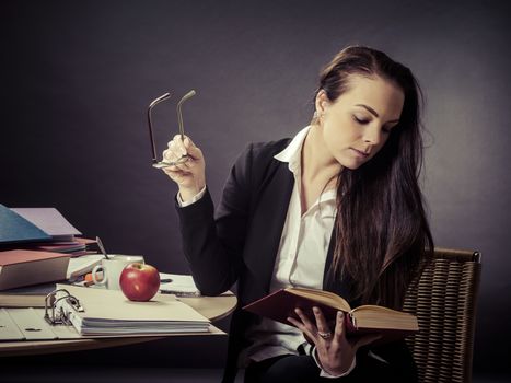 Photo of a teacher in her 30's sitting at a messy desk in front of a large blackboard reading a book.