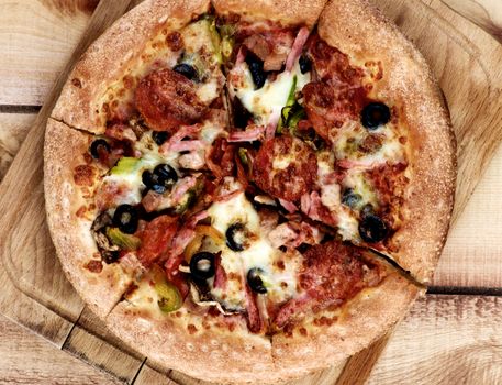 Homemade Fresh Pepperoni Pizza with Black Olives, Ham, Cheese and Tomatoes on Cutting Board closeup on Wooden background. Top View