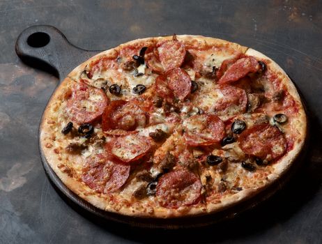 Freshly Baked Homemade Meat Pizza with Salami, Meat Balls, Ham, Black Olives and Mozarella Cheese on Wooden Cutting Board closeup on Grange Dark background