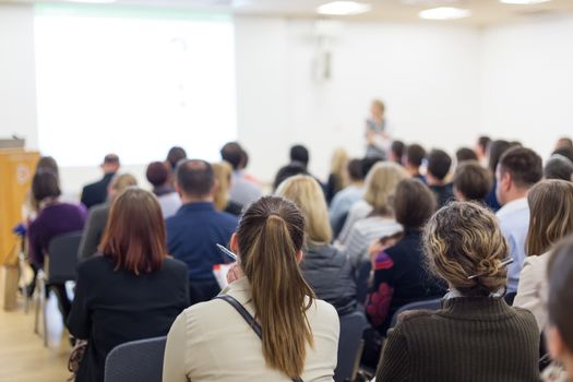 Business and entrepreneurship symposium. Female speaker giving a talk at business meeting. Audience in conference hall. Rear view of unrecognized participant in audience. Copy space on whitescreen.