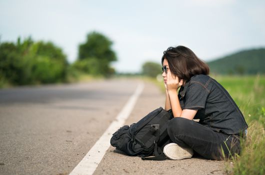 Young asian woman short hair and wearing sunglasses sit with backpack hitchhiking along a road wait for help in country road Thailand