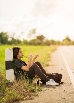Young asian woman short hair and wearing sunglasses sit with backpack hitchhiking along a road wait for help in country road Thailand