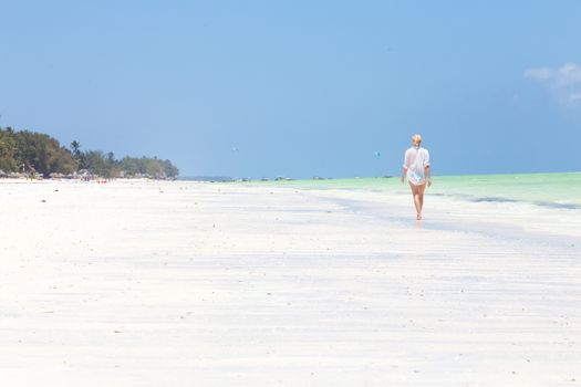 Happy woman having fun, enjoying summer, walking along white tropical beach. Beautiful caucasian model wearing white beach tunic on vacations on picture perfect Paje beach, Zanzibar, Tanzania.